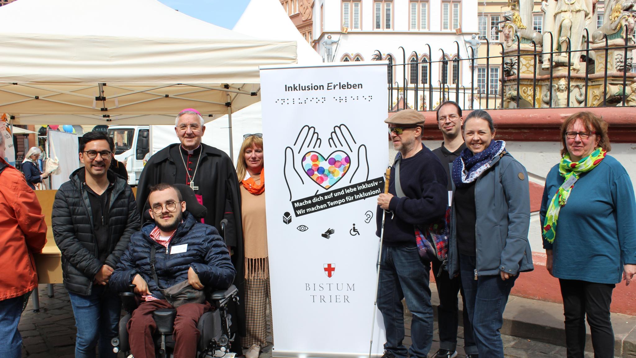 Menschen mit und ohne Behinderung stehen auf dem Hauptmarkt in Trier vor einem Aktionszelt während der Heilig-Rock-Tage 2022. In ihrer Mitte ist ein hohes Plakat zu sehen. Darauf steht oben 'Inklusion Erleben' und unten 'Bistum Trier'. Dazwischen sind die Umrisse von zwei Händen aufgedruckt, die ein Herz halten, das mit bunten Punkten gefüllt ist. Zur Gruppe gehören Weihbischof Franz-Josef Gebert, Mitarbeiterinnen und Mitarbeiter der Tonpost und aus dem Team Inklusion.