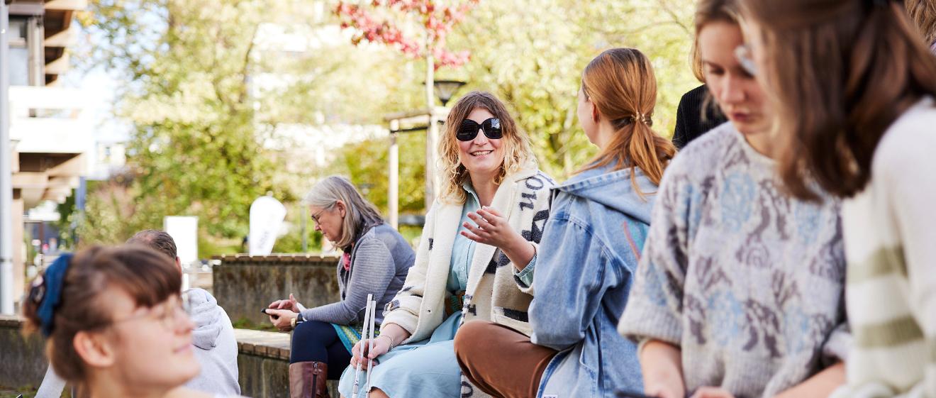 Mehrere Frauen sitzen auf stufenförmig angeordneten Holzbänken. Im Vordergrund drei Personen, die sich unterhalten und lächeln. Im Hintergrund zwei Frauen, die sich unterhalten, eine davon trägt eine schwarze Brille und einen Langstock.