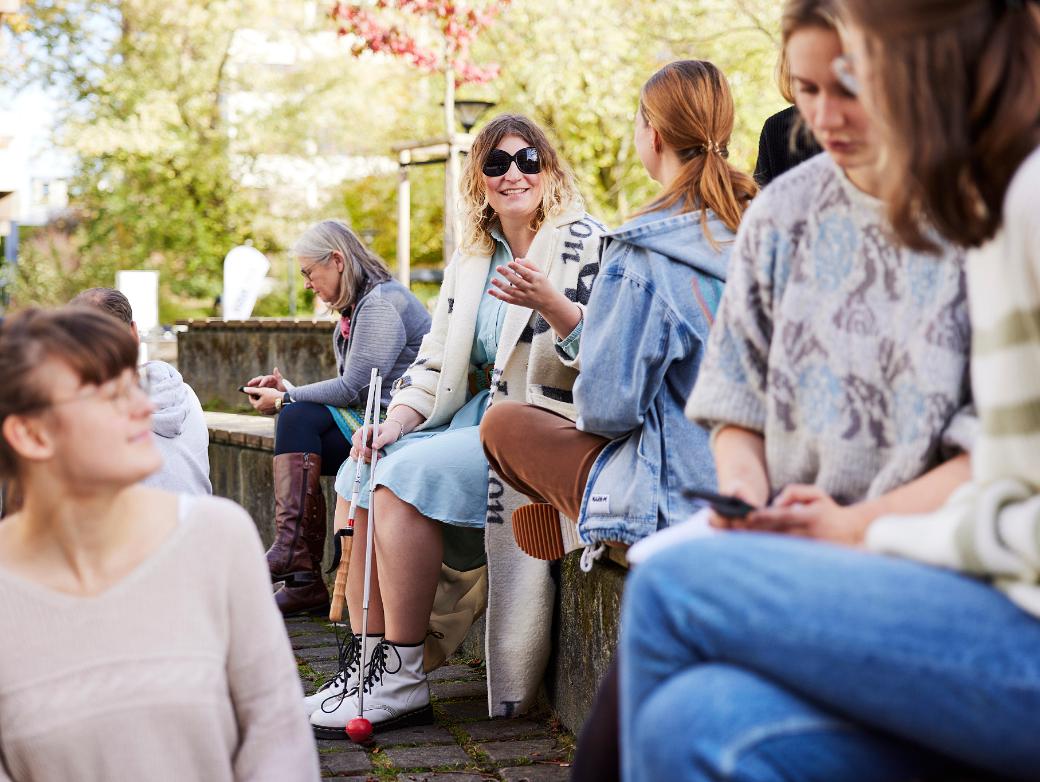 Mehrere Frauen sitzen auf stufenförmig angeordneten Holzbänken. Im Vordergrund drei Personen, die sich unterhalten und lächeln. Im Hintergrund zwei Frauen, die sich unterhalten, eine davon trägt eine schwarze Brille und einen Langstock.