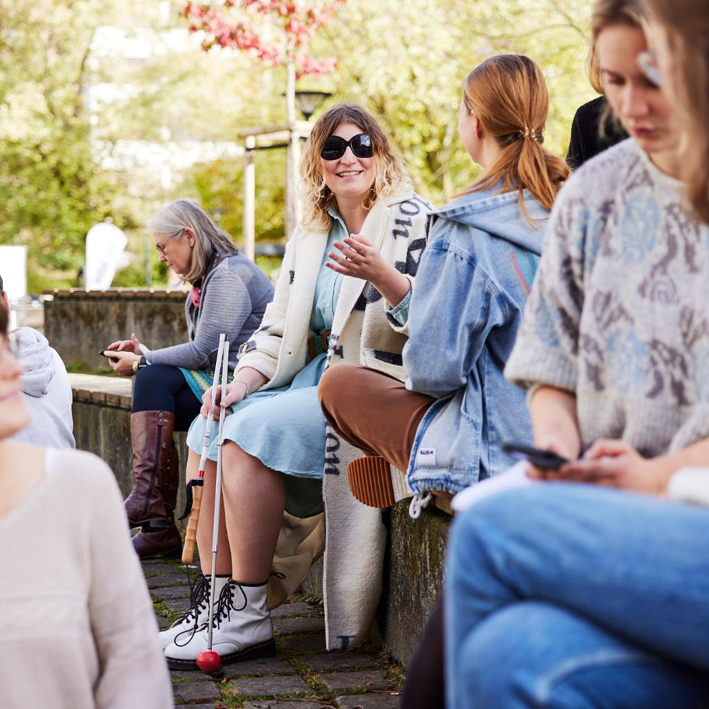 Mehrere Frauen sitzen auf stufenförmig angeordneten Holzbänken. Im Vordergrund drei Personen, die sich unterhalten und lächeln. Im Hintergrund zwei Frauen, die sich unterhalten, eine davon trägt eine schwarze Brille und einen Langstock.