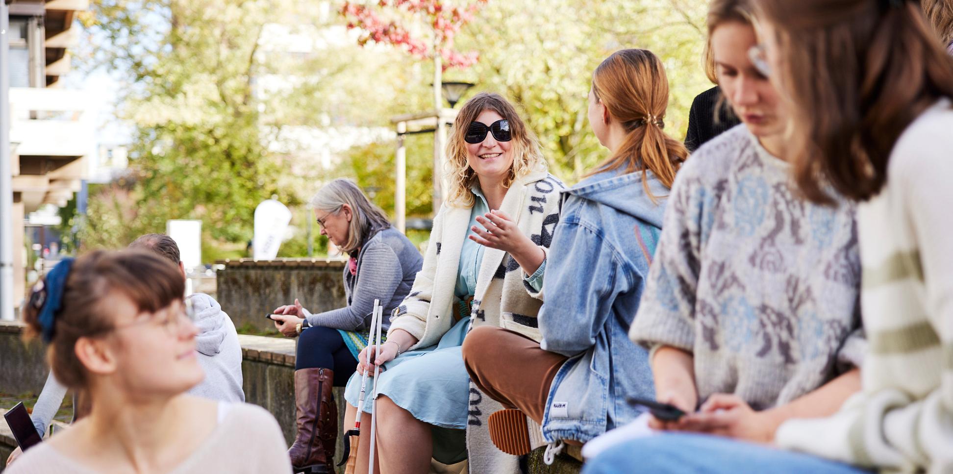 Mehrere Frauen sitzen auf stufenförmig angeordneten Holzbänken. Im Vordergrund drei Personen, die sich unterhalten und lächeln. Im Hintergrund zwei Frauen, die sich unterhalten, eine davon trägt eine schwarze Brille und einen Langstock.
