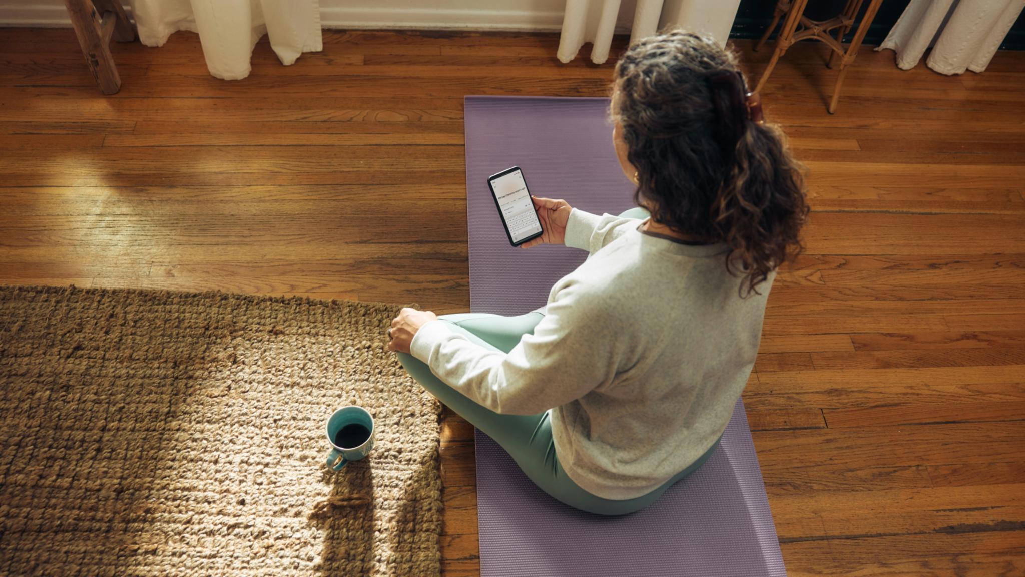 Frau im weißen Shirt sitzt auf Yogamatte mit Smartphone in der Hand und Kaffeetasse