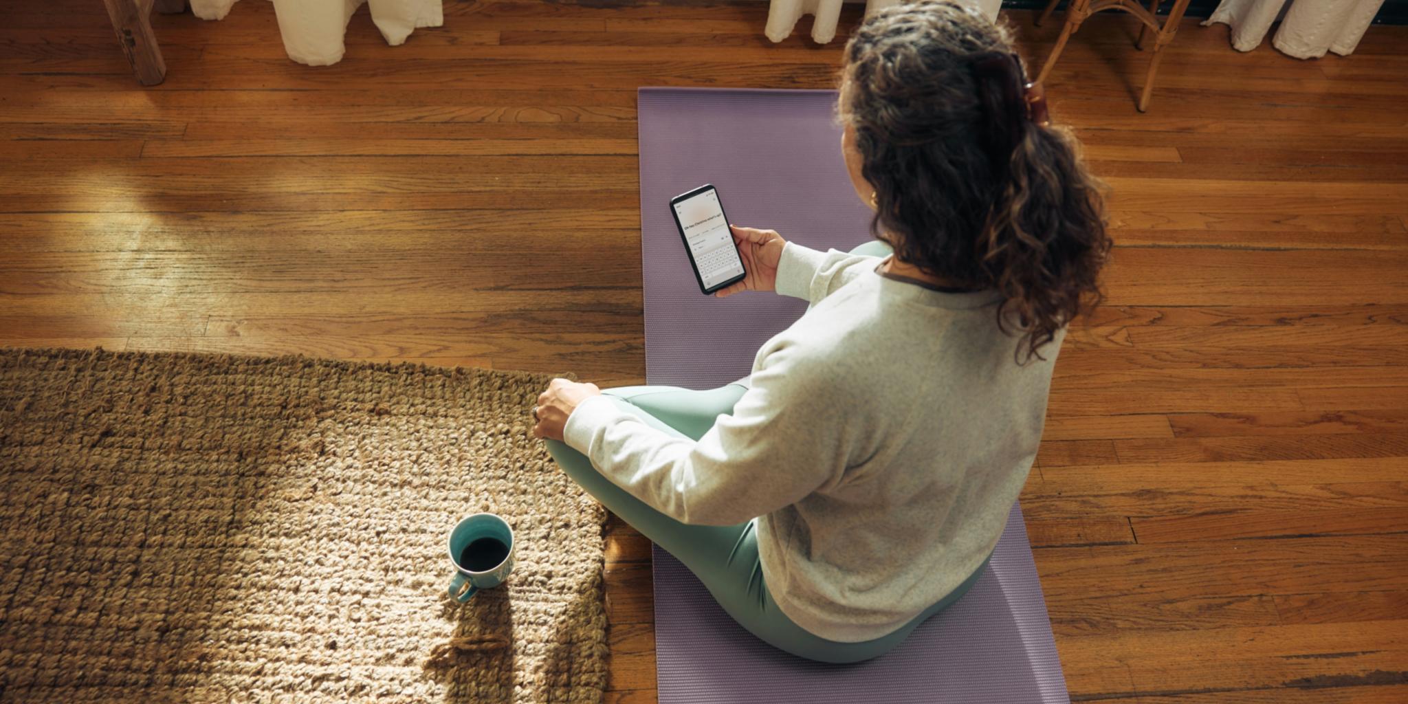 Frau im weißen Shirt sitzt auf Yogamatte mit Smartphone in der Hand und Kaffeetasse